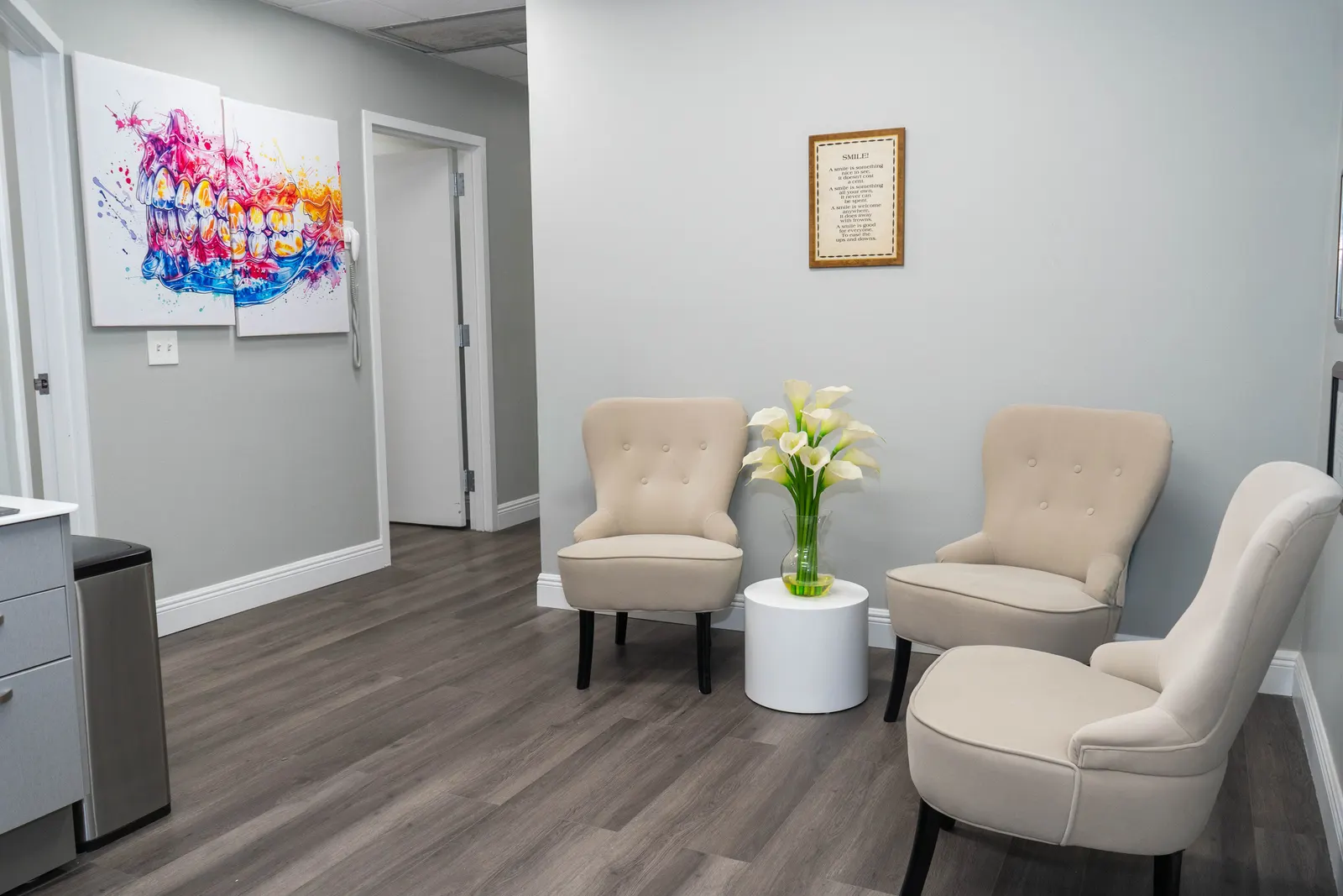 Waiting room with three beige chairs, a white side table with flowers, and colorful dental art on wall.