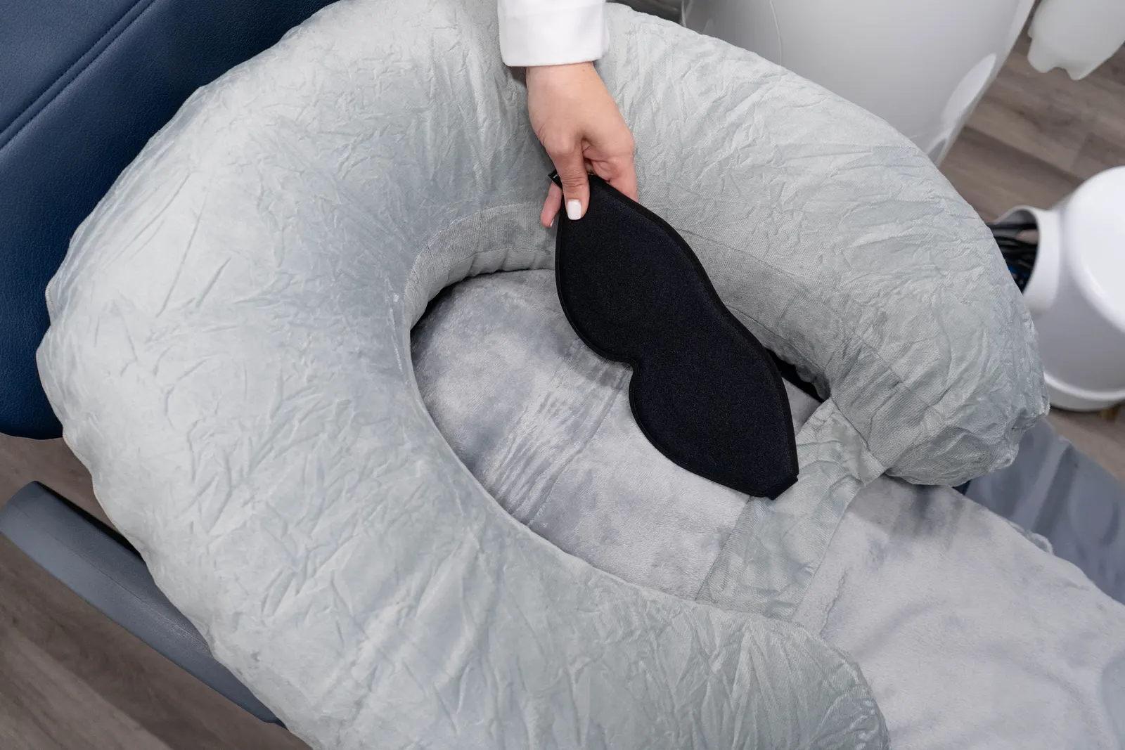 Hand placing a black eye mask on a gray padded headrest of a medical or spa chair.