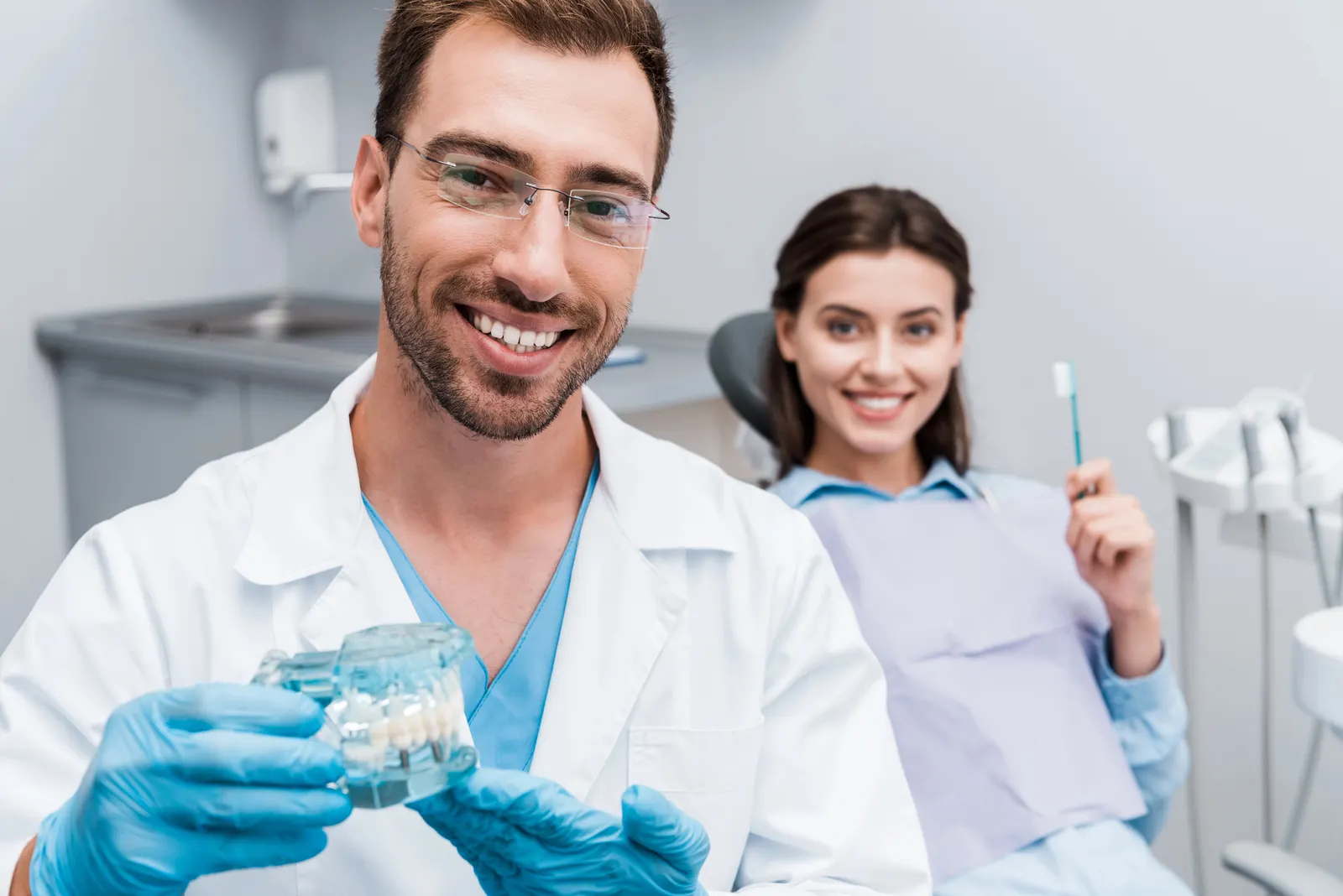 Smiling dentist holding a dental model with a patient holding a toothbrush behind him in clinic.