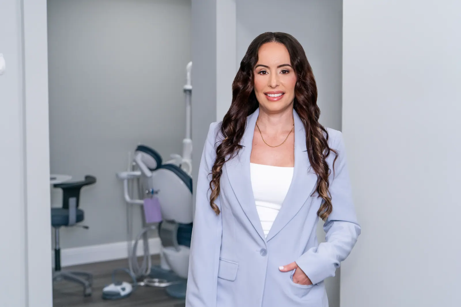 Smiling woman with long wavy hair wearing a pale blue blazer in a dental office.