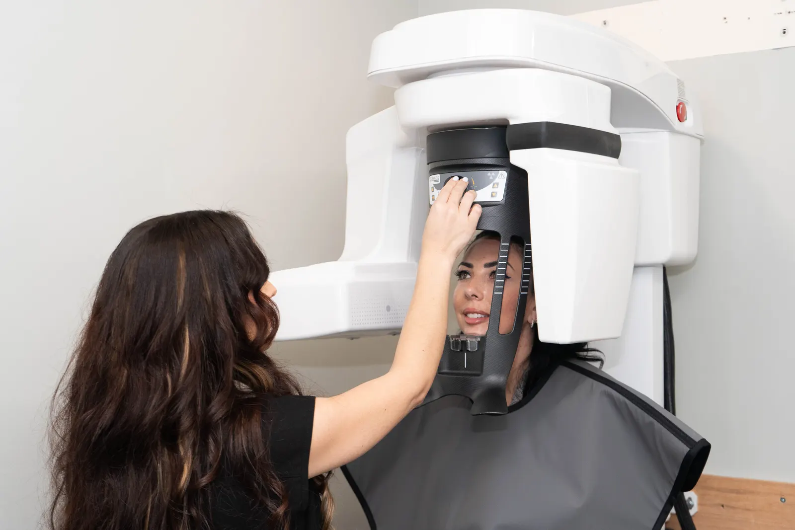 Technician adjusts a dental X-ray machine while a patient wears a protective lead apron.