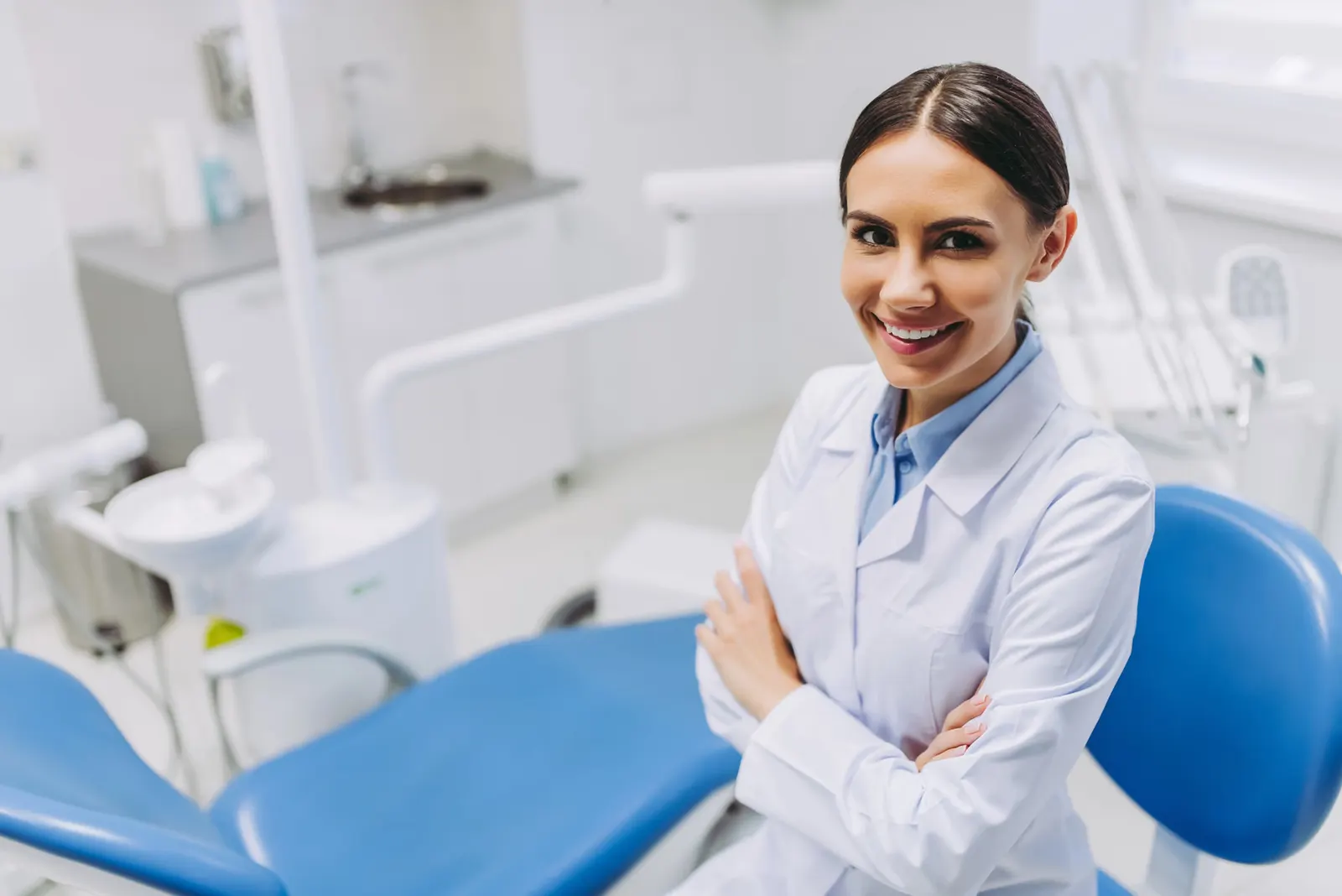 Smiling female dentist in white coat standing with arms crossed in dental clinic with blue chair.