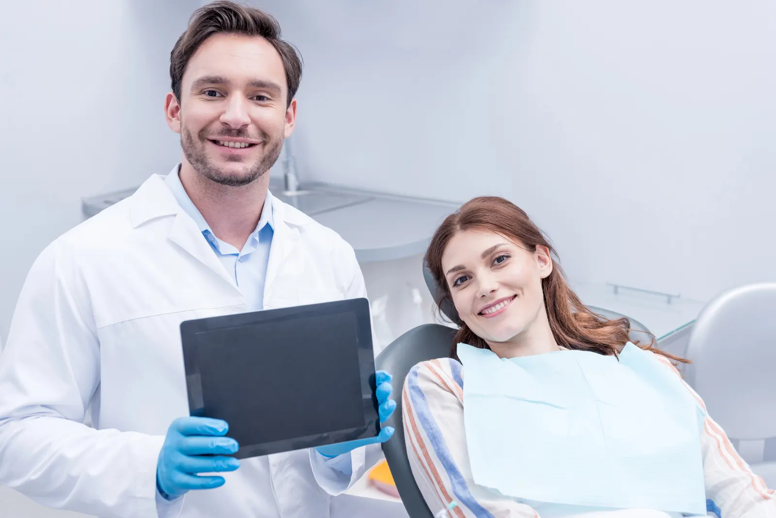Smiling male dentist in gloves holding a tablet next to a female patient in a dental chair.