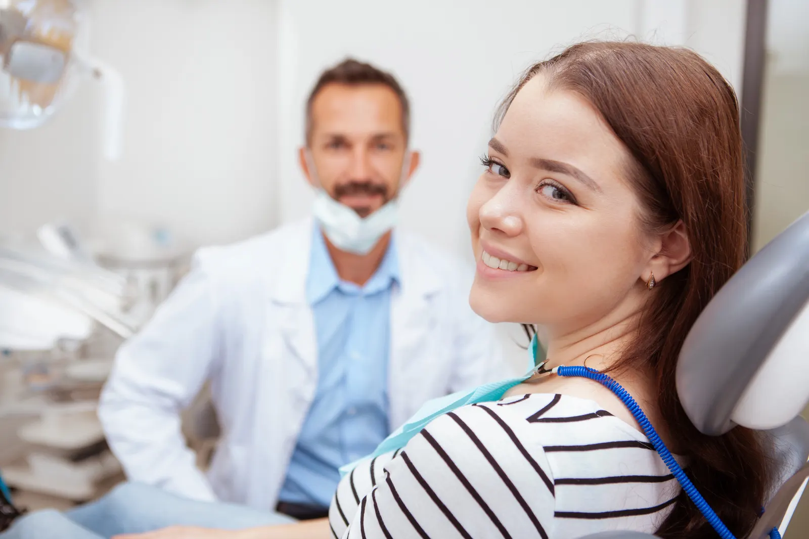 Smiling young woman sitting in dental chair with dentist in background wearing a mask.