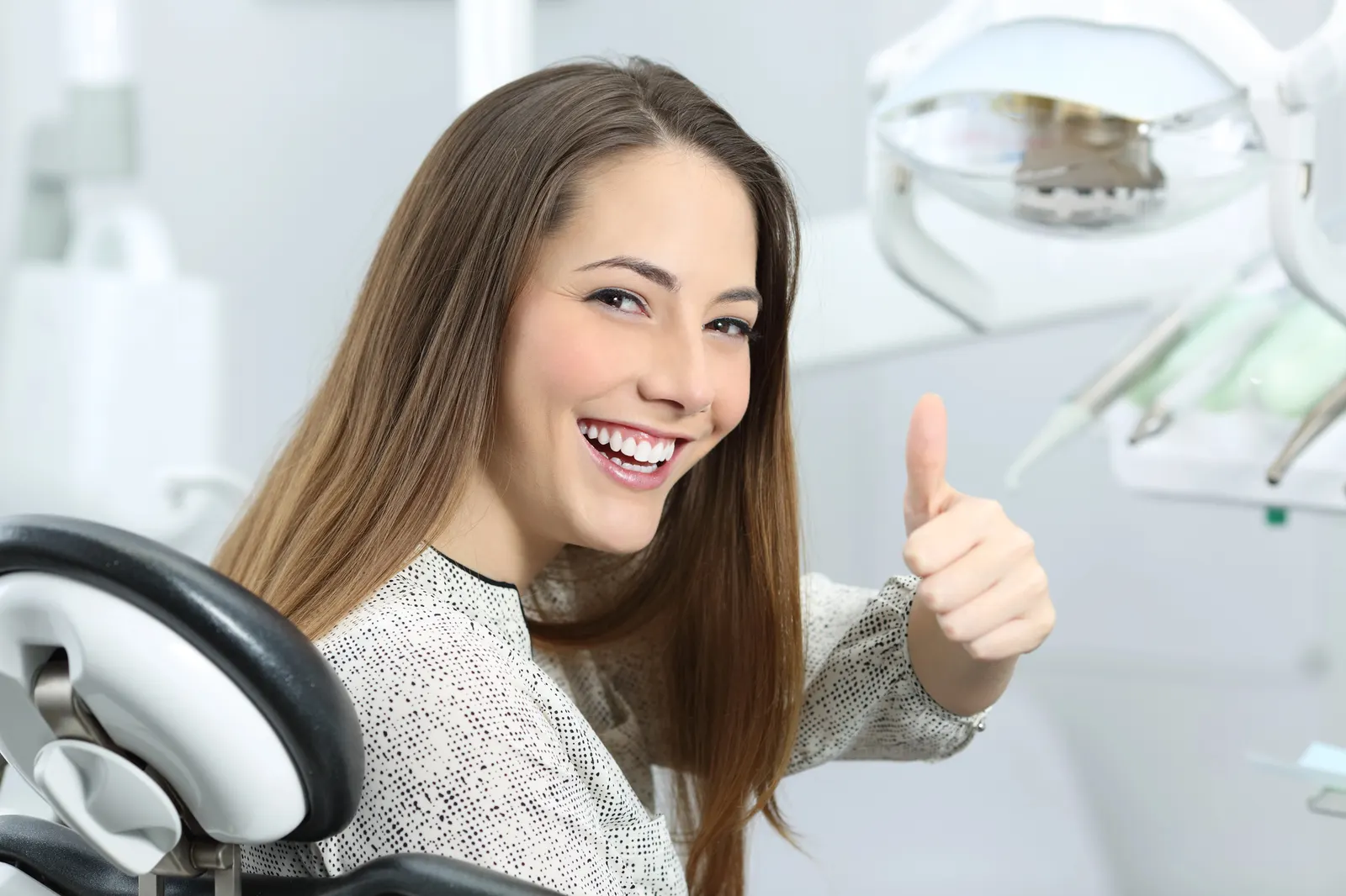 Smiling woman in dental chair giving thumbs-up with dental equipment in background.