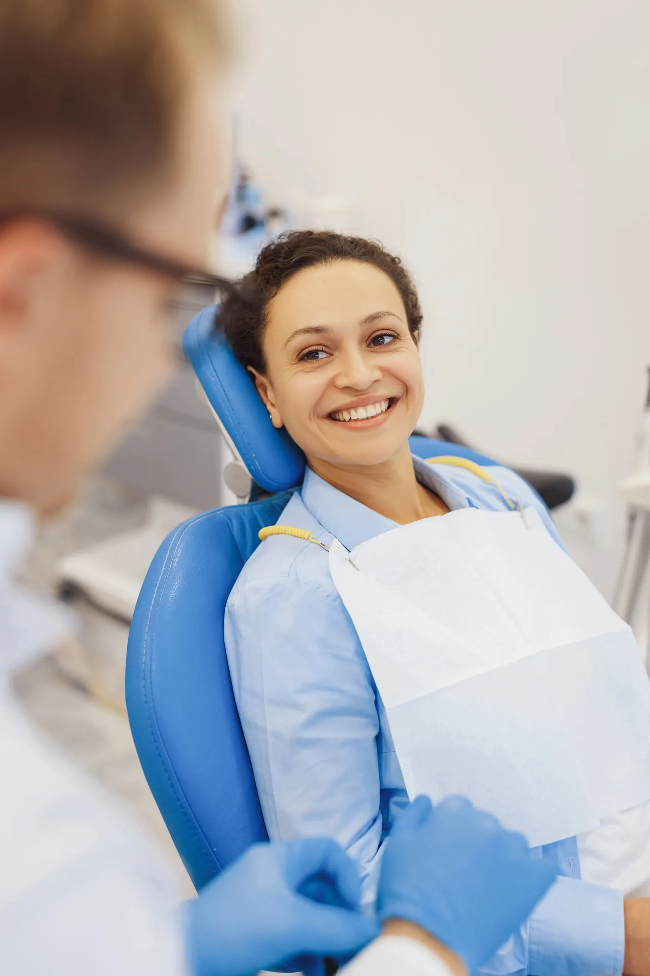 Smiling woman in dental chair wearing bib as dentist prepares tools wearing blue gloves.