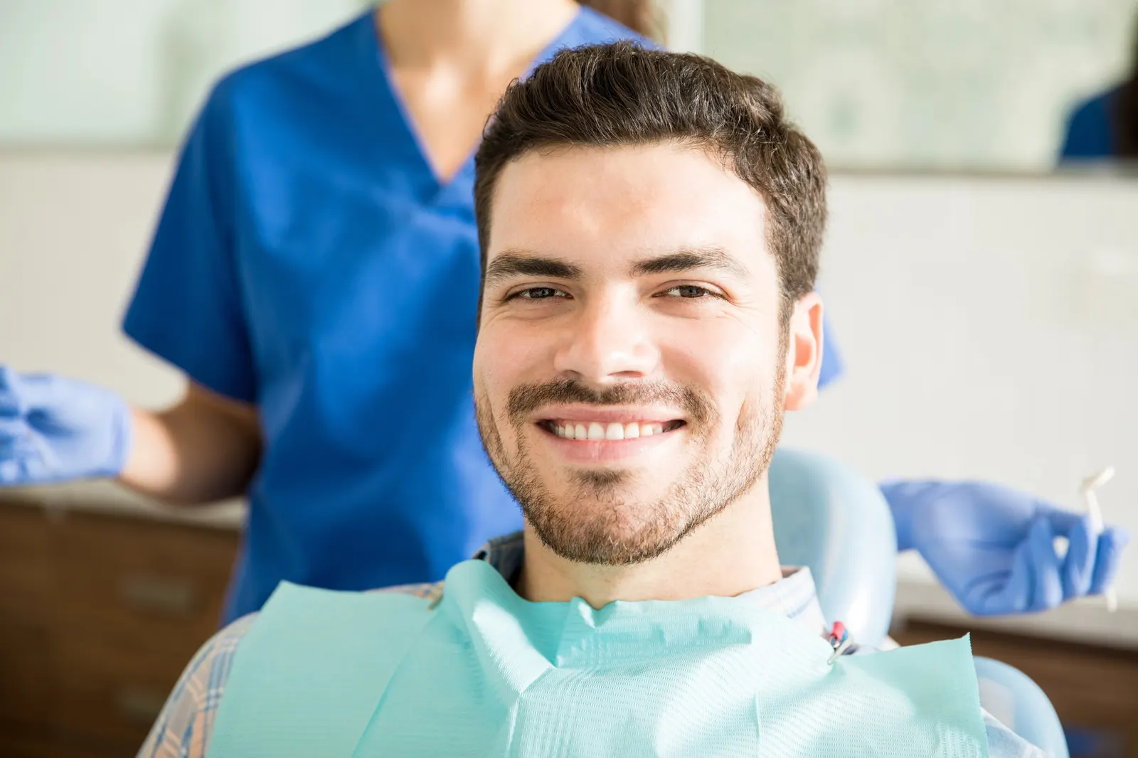 Smiling man in a dental chair with a dental hygienist holding tools in the background.