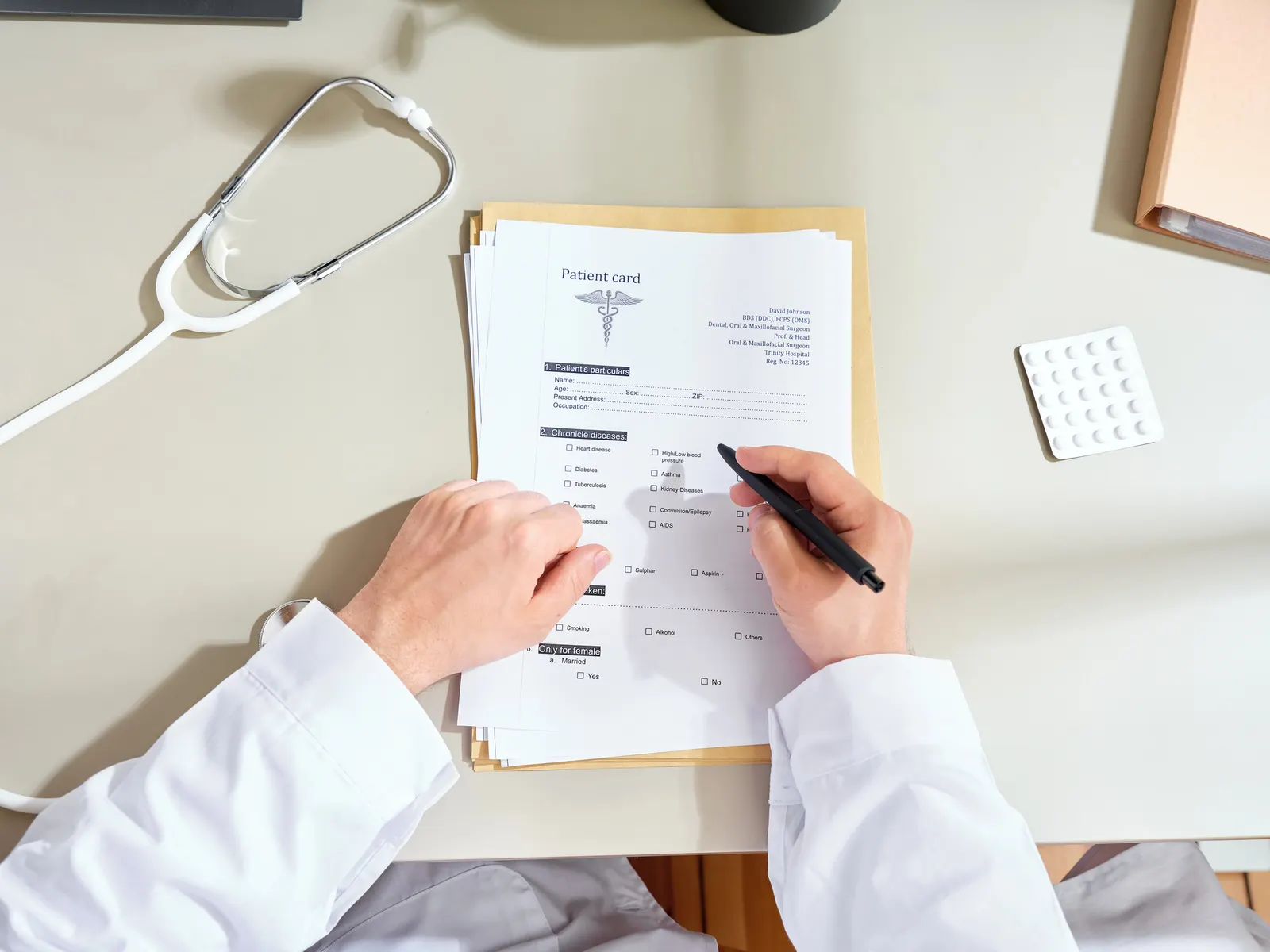 Doctor filling out patient card form on desk with stethoscope and medication nearby.