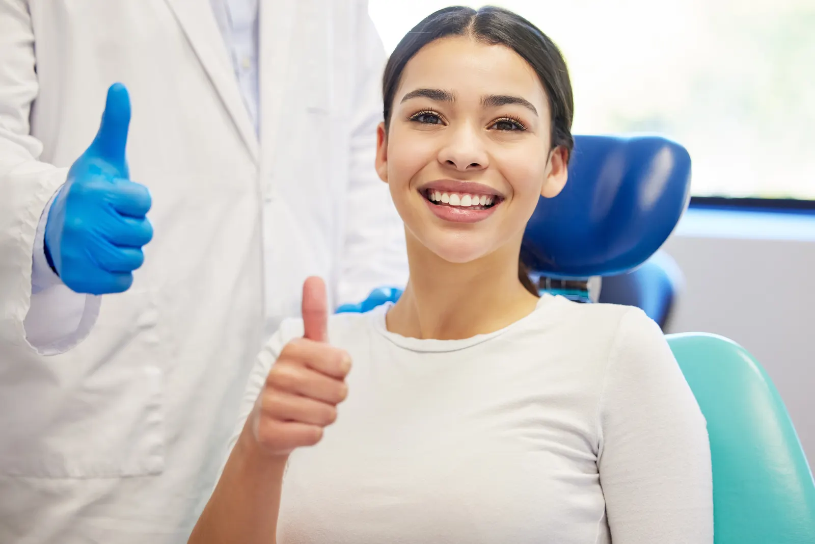 Smiling woman in dental chair with dentist giving thumbs up wearing blue gloves.