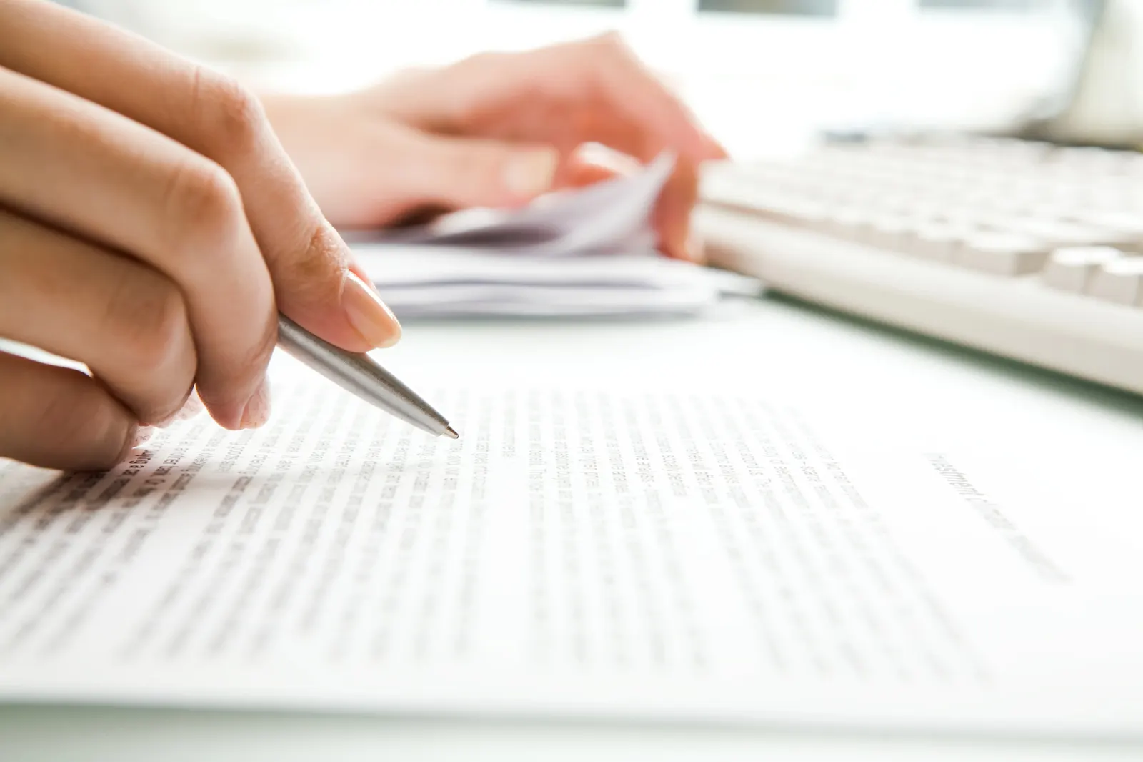 Hand holding pen pointing at text on paper with keyboard in background.