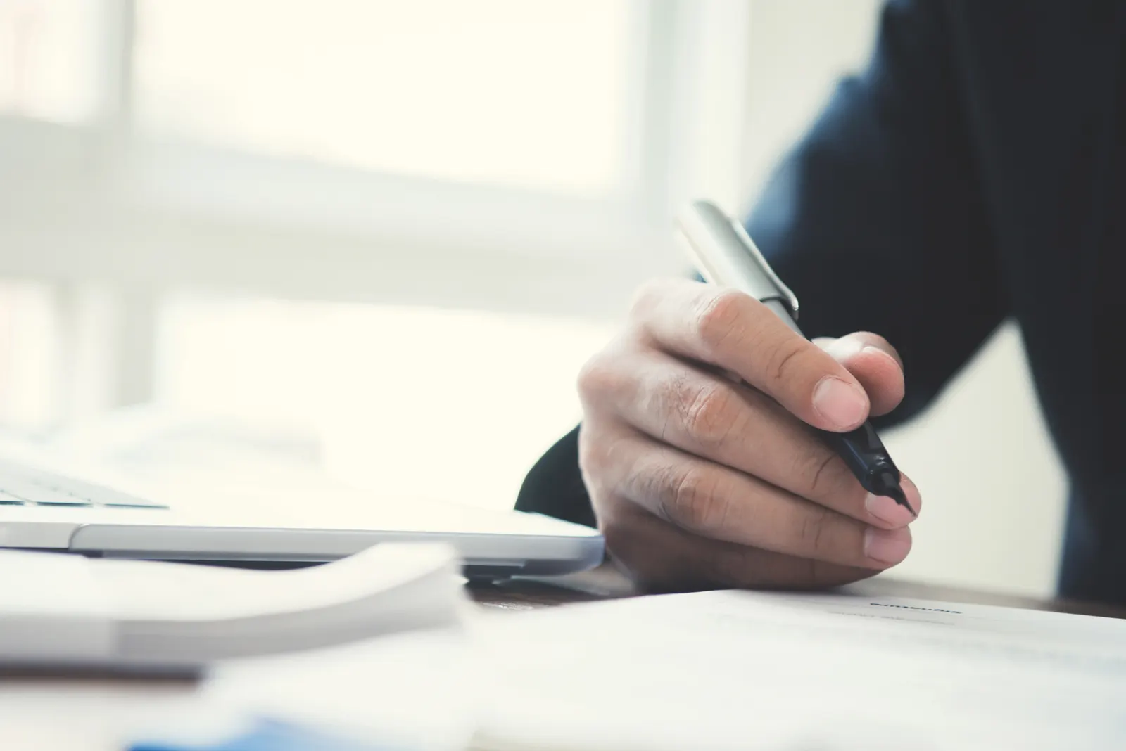 Person's hand holding a pen over documents with a laptop keyboard visible on the desk.