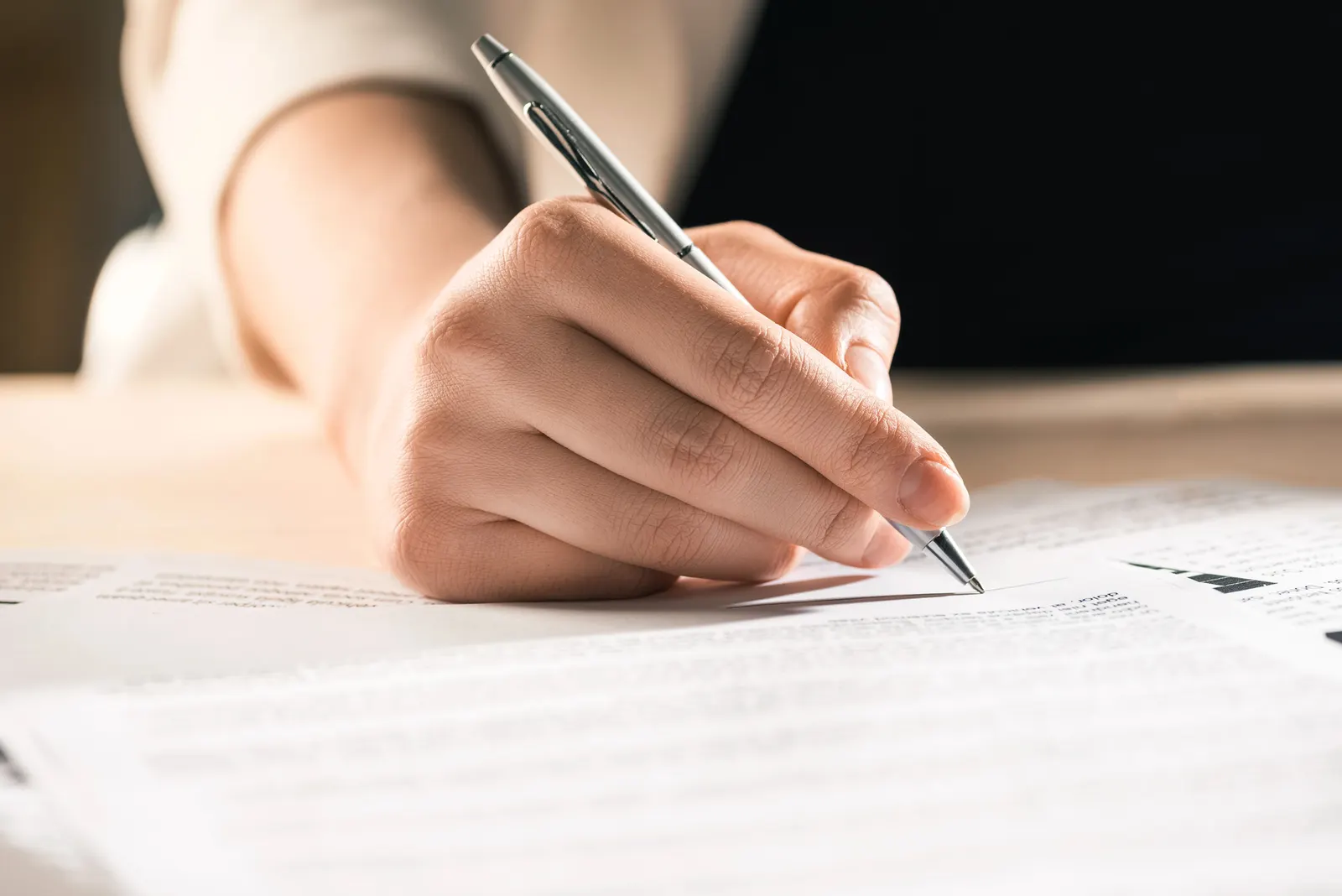 Person signing a document with a silver pen on a wooden table with blurred text papers.