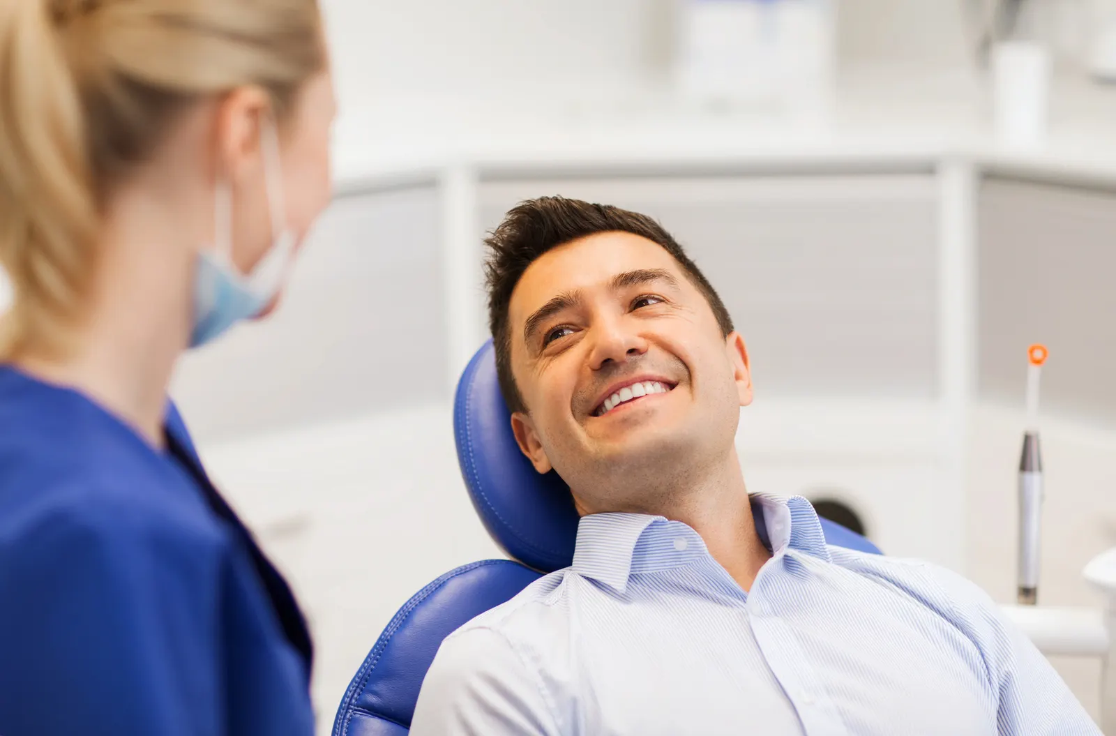Smiling man reclined in a dental chair looking at a female dentist wearing a mask and blue scrubs.