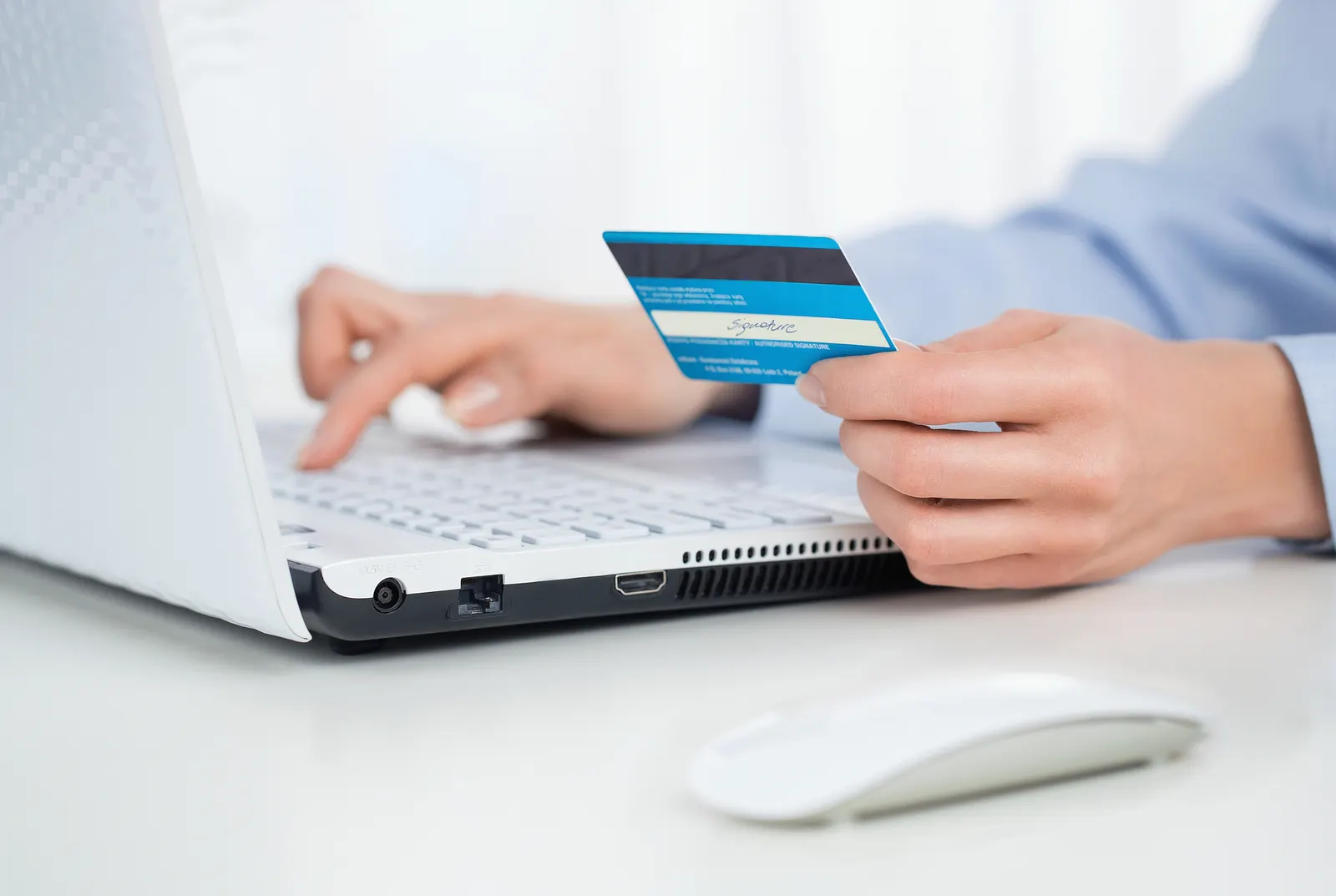Person holding credit card and typing on a white laptop keyboard with a wireless mouse nearby.
