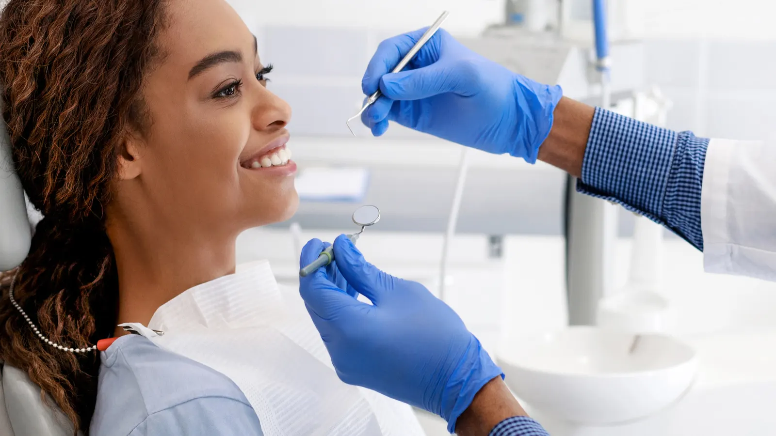 A smiling patient in a dental chair being examined by a dentist wearing blue gloves holding dental tools.