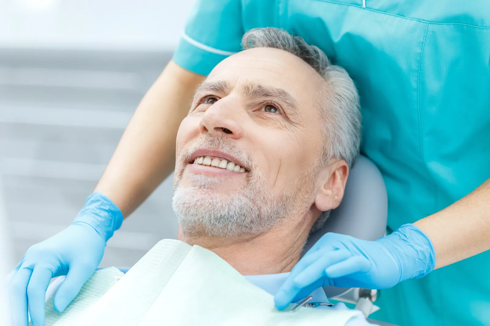 Smiling older man reclined in dental chair with gloved hands of dental professional nearby.