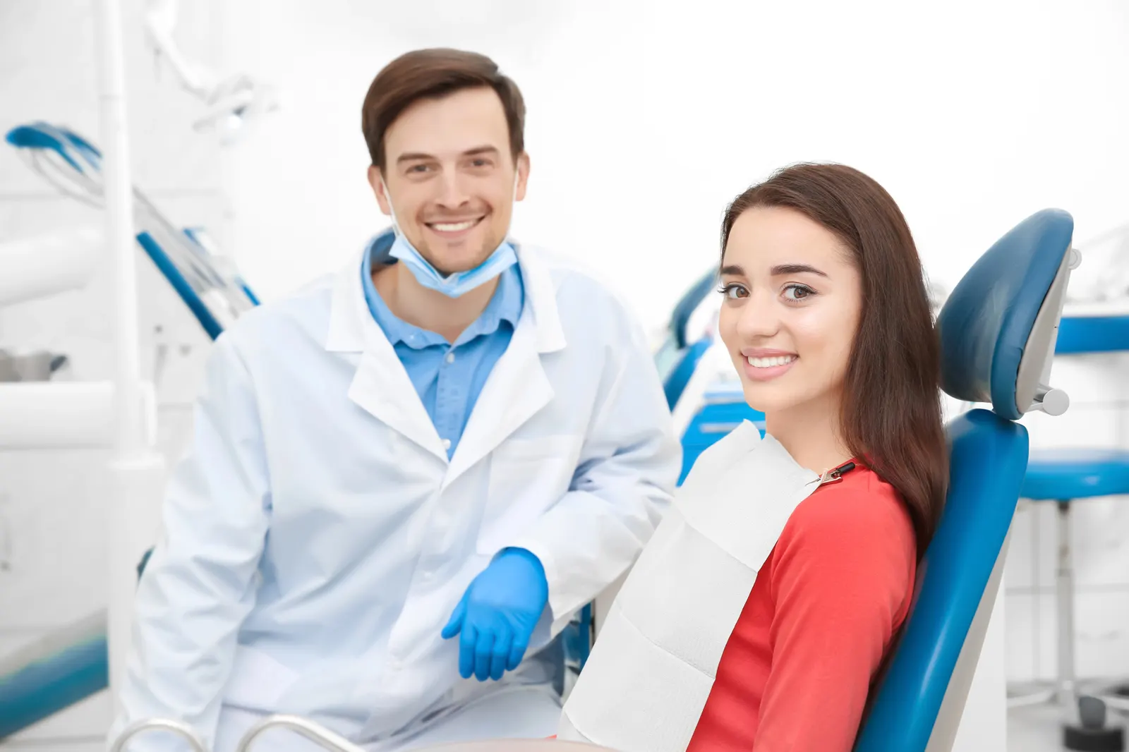 Smiling dentist in white coat and blue gloves with female patient sitting in dental chair wearing bib.