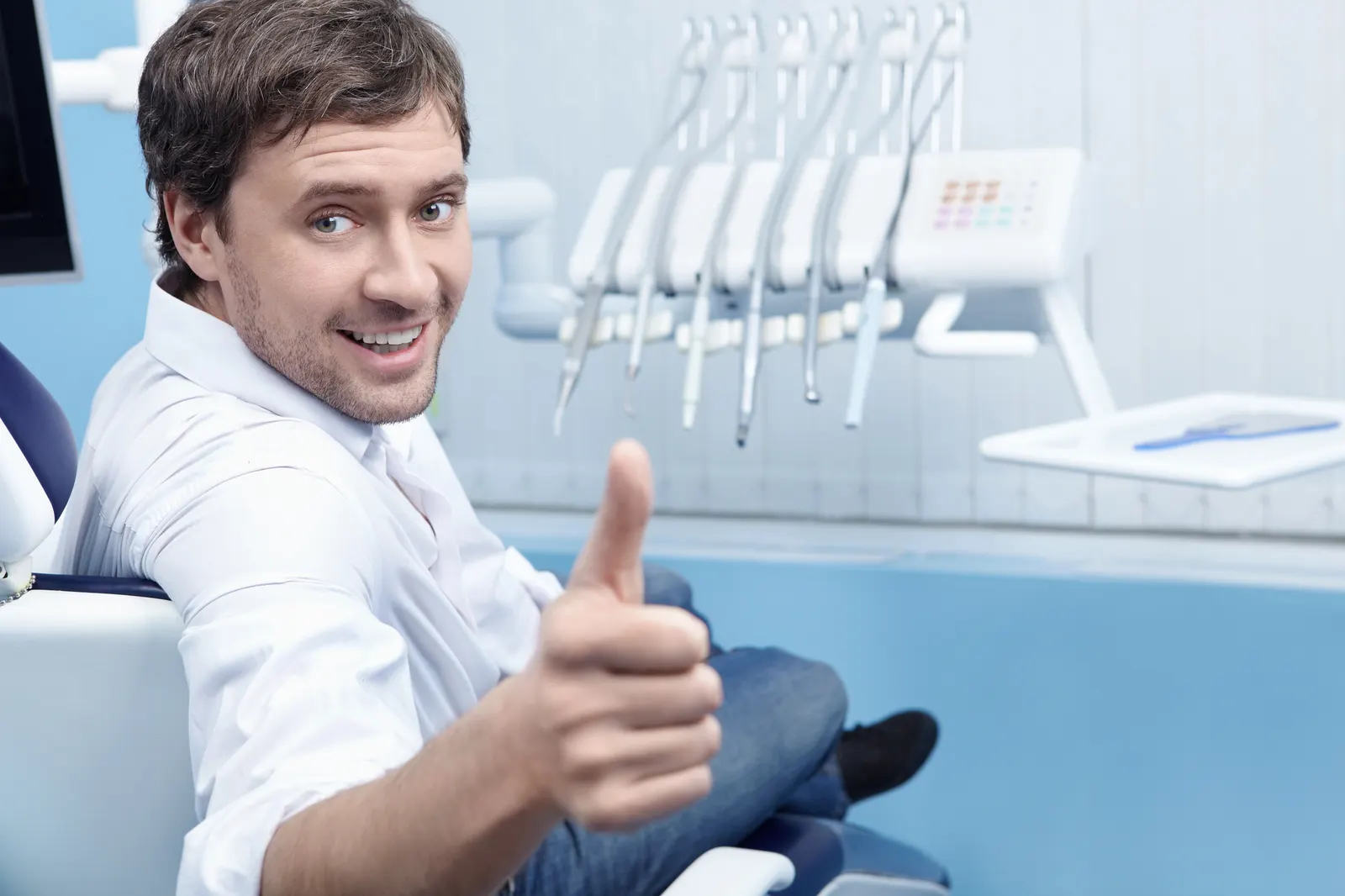 Smiling man in dental chair giving a thumbs-up with dental tools in the background.