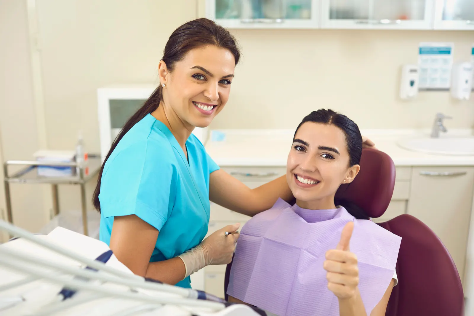 Female dentist smiling beside a smiling patient giving thumbs up in a dental office.
