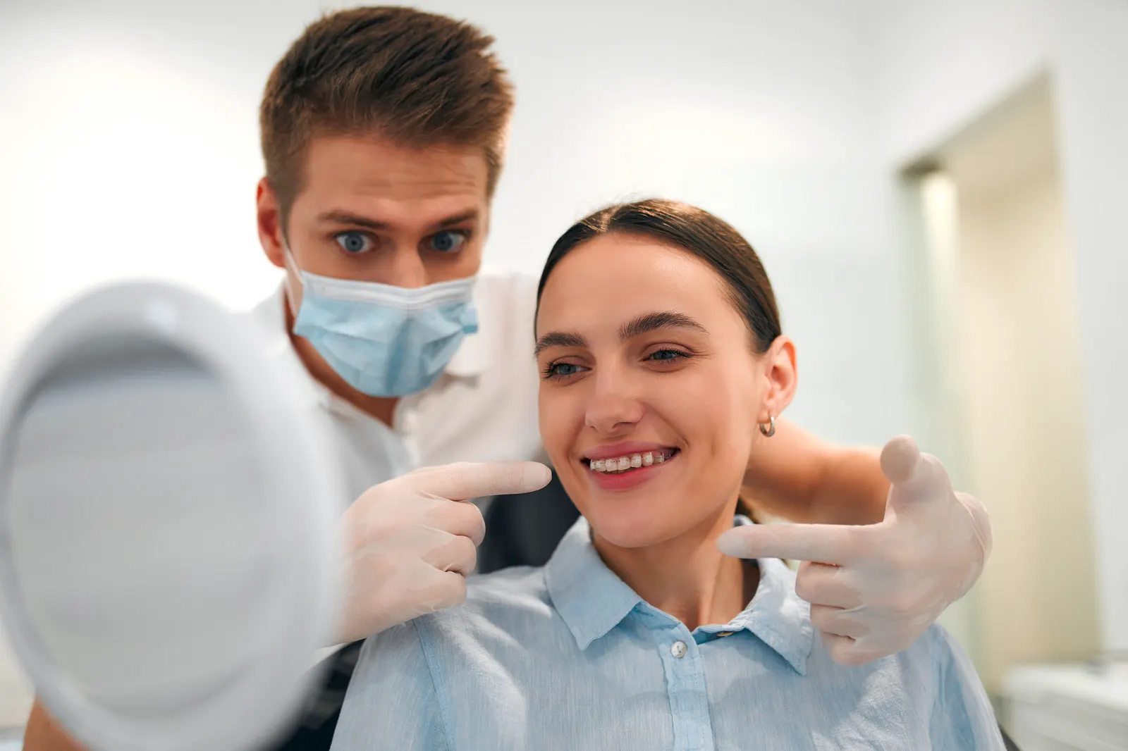 Dentist with mask and gloves pointing at a smiling woman’s teeth with clear braces in dental office.