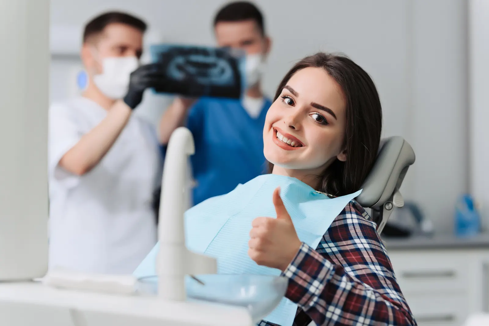 Smiling woman in dental chair giving thumbs up, while dentists with masks review X-ray in background.