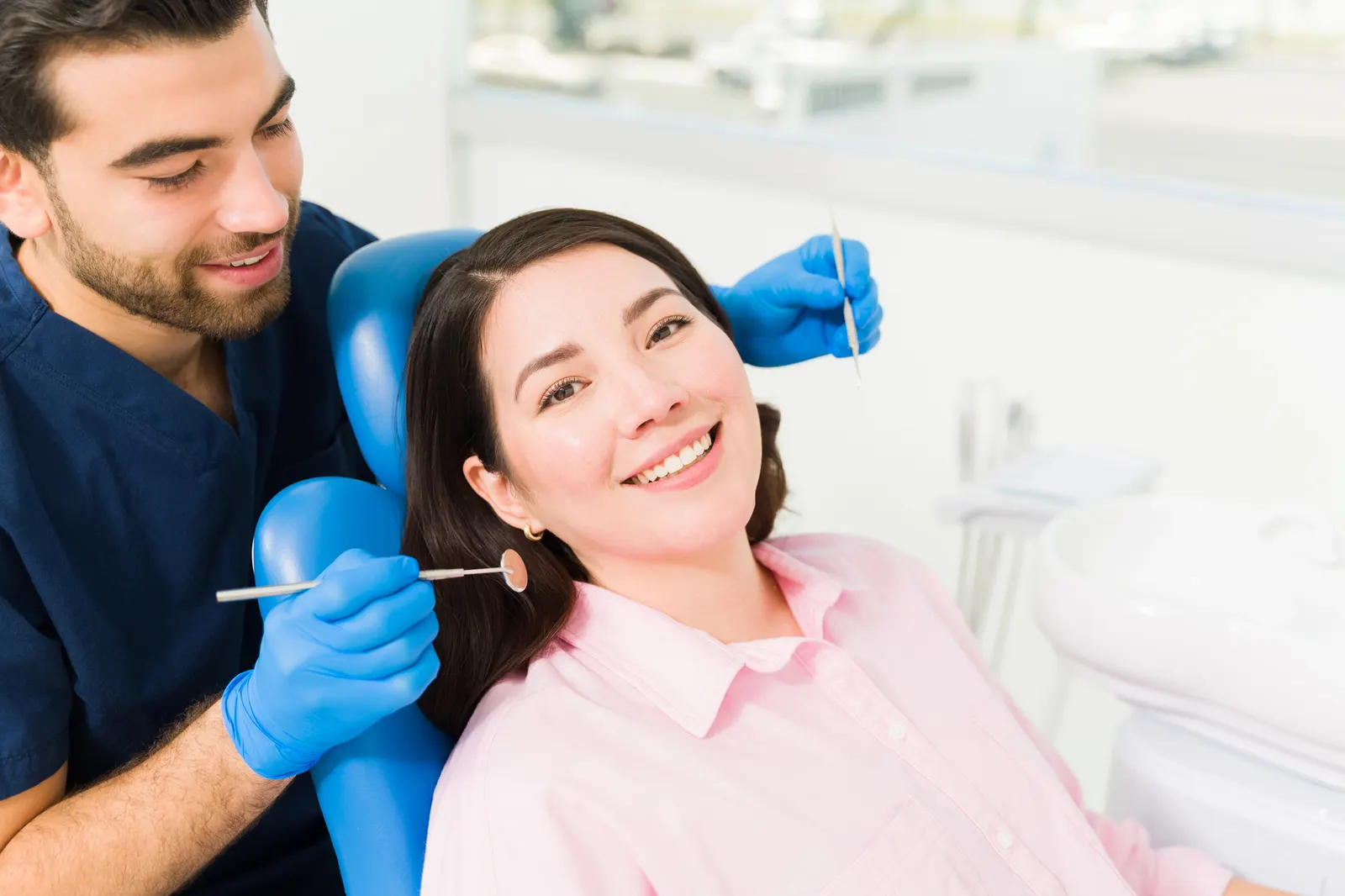 Dentist wearing blue gloves examining smiling woman's teeth in dental chair.