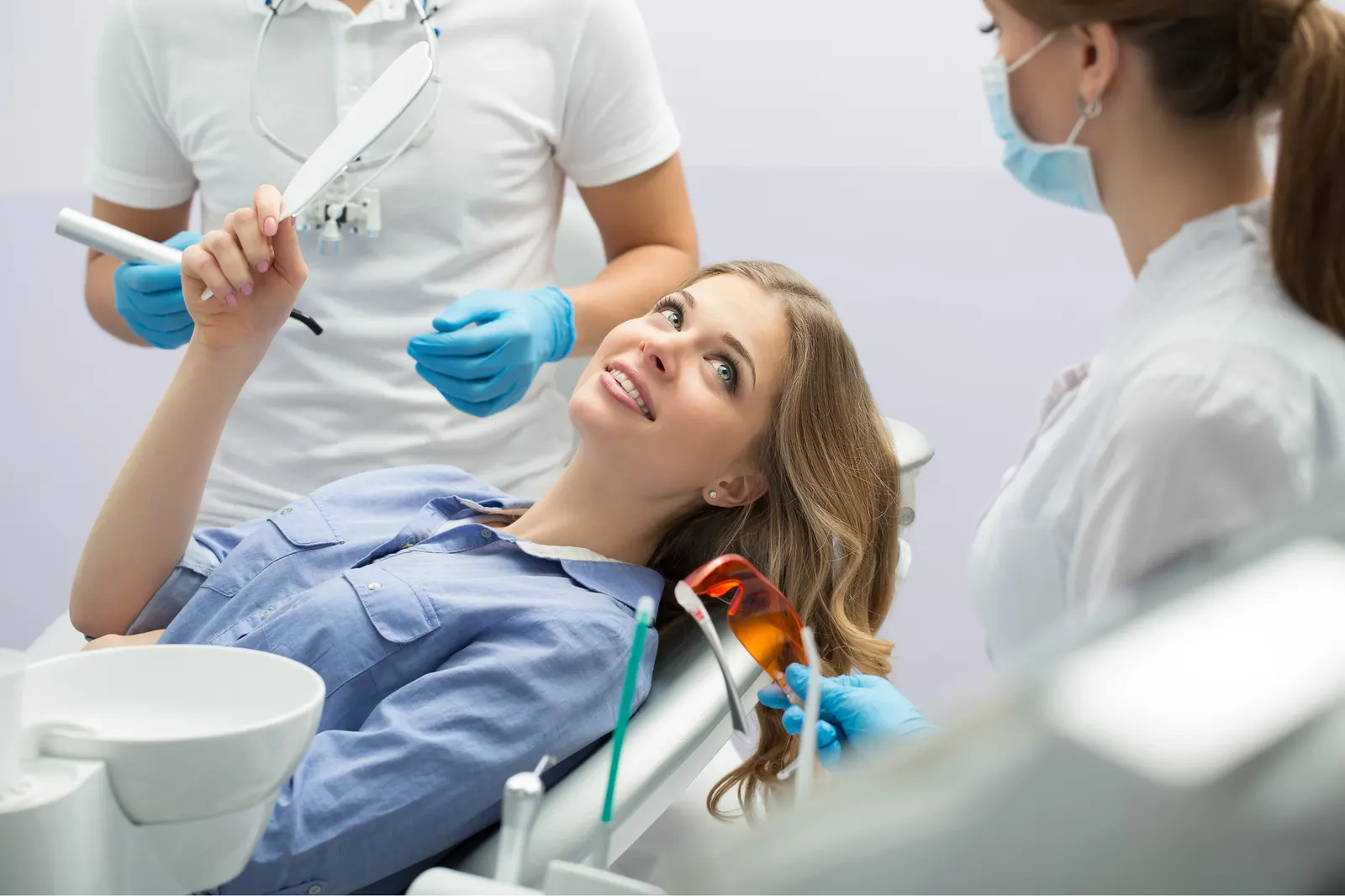 Woman in dental chair holding a mirror looking at dentist and assistant wearing masks and gloves.