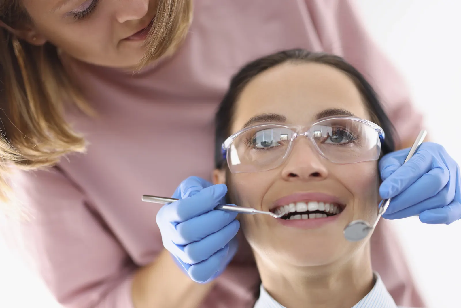 Dentist wearing gloves examines smiling patient's teeth using dental mirror and probe.