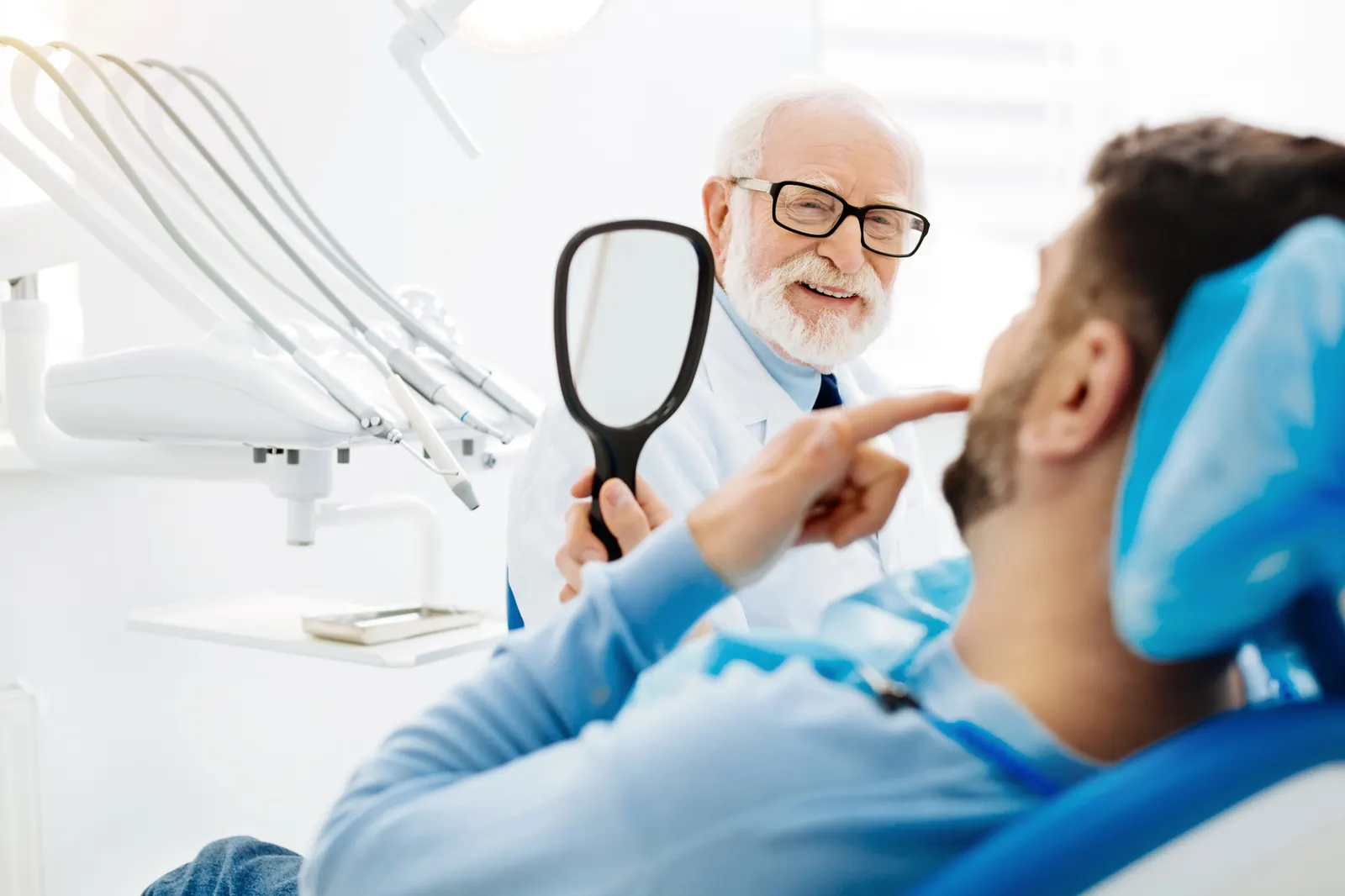 Elderly male dentist in glasses smiling and holding a mirror for a seated male patient in dental chair.
