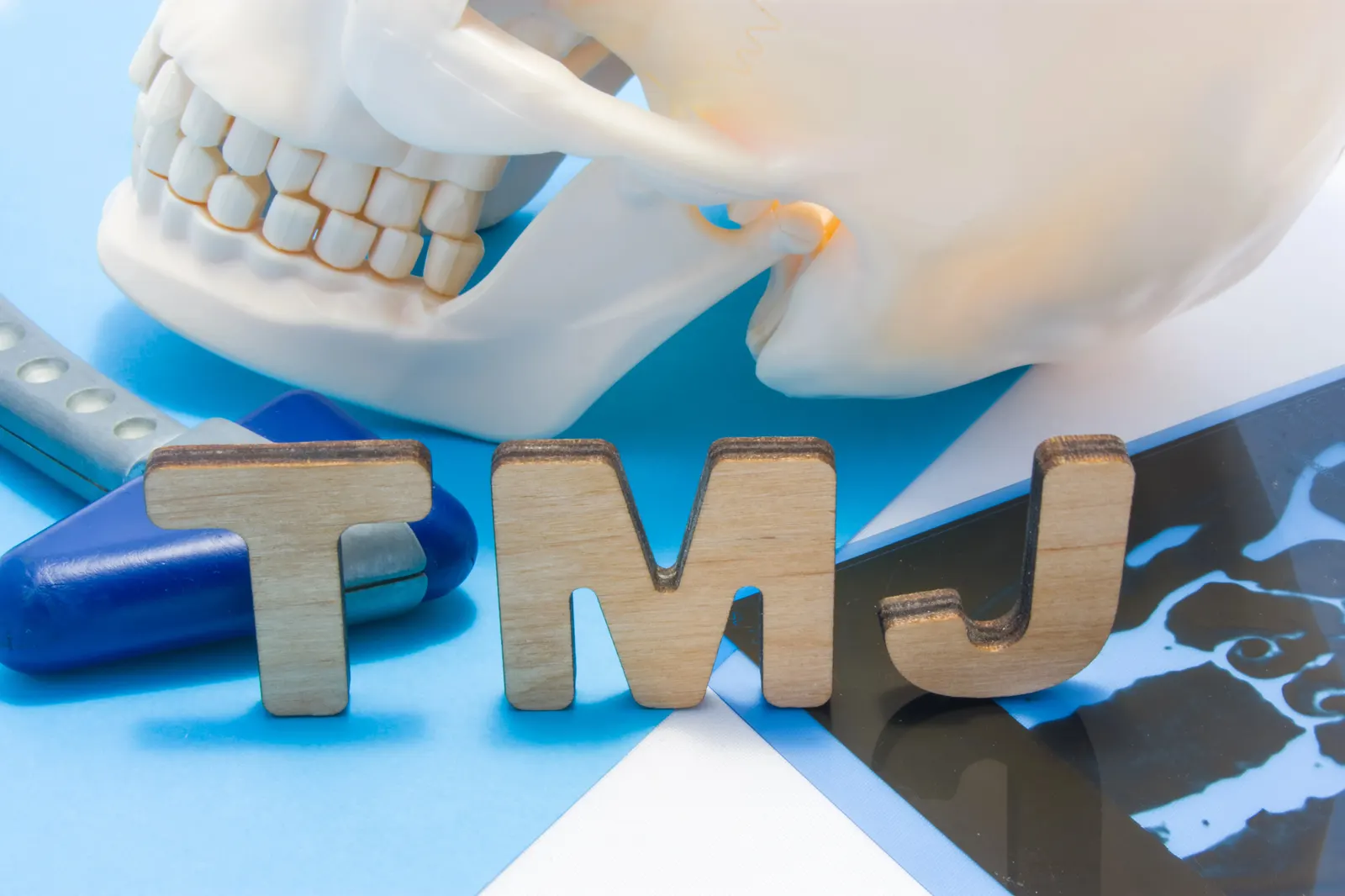 Wooden letters T M J with a skull model and jaw adjusting tool on a blue background.