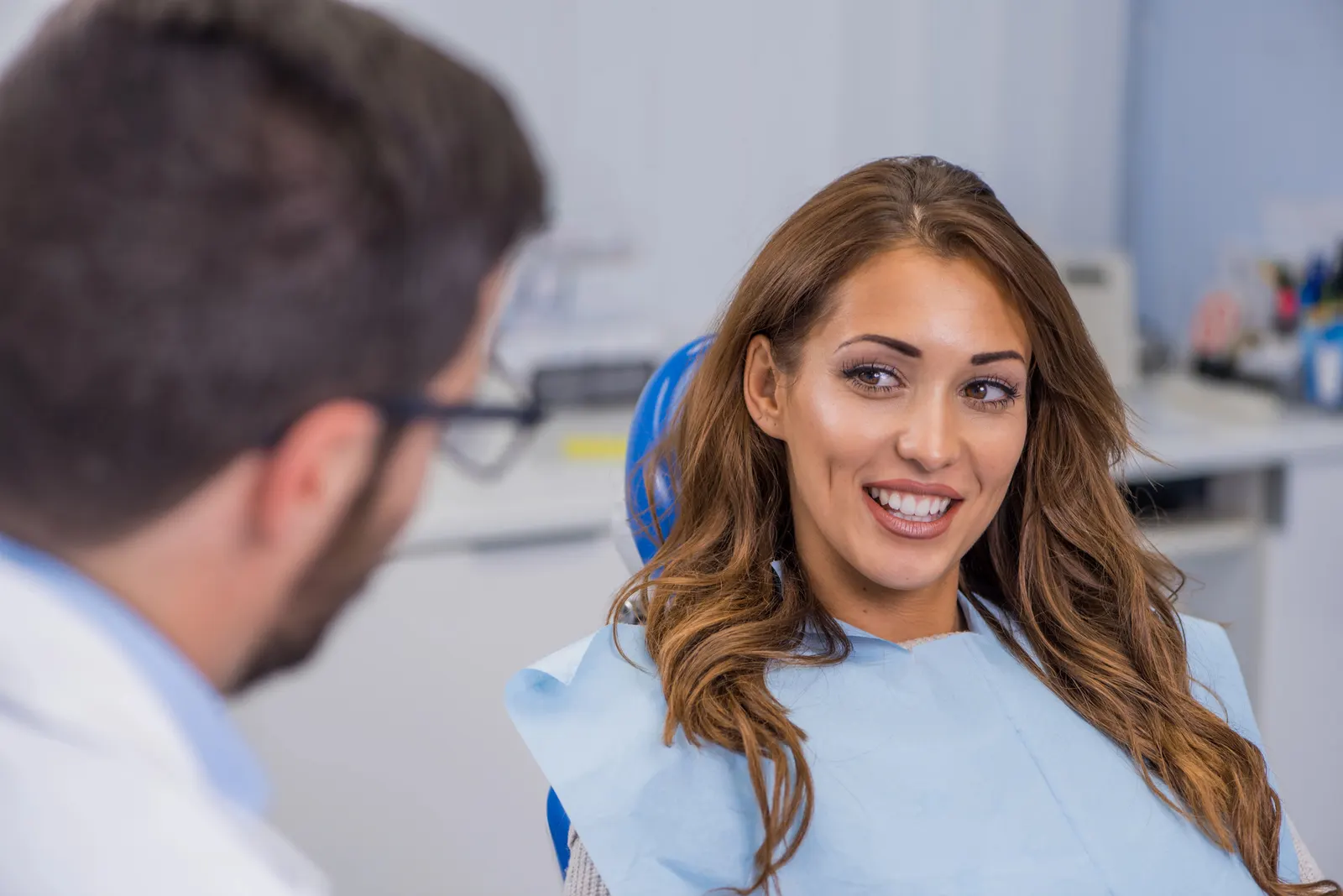 Smiling woman sitting in dental chair talking to male dentist wearing glasses and white coat.