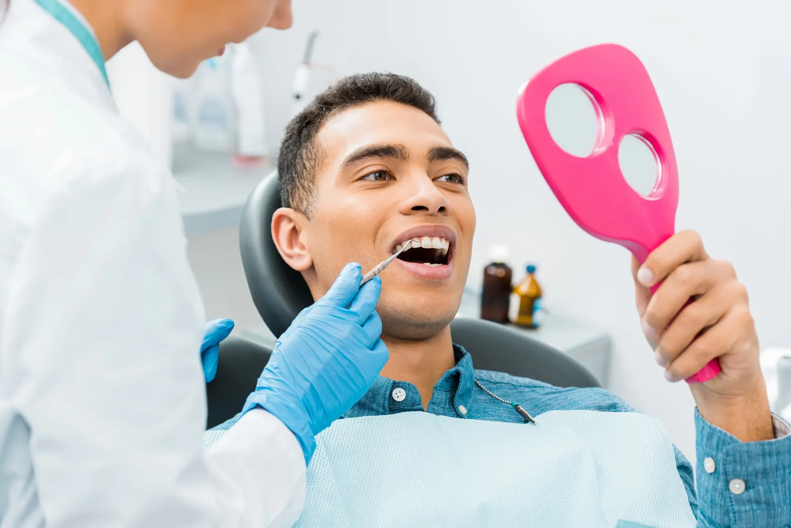Man at dental checkup holding pink mirror while dentist examines his teeth with dental tool.