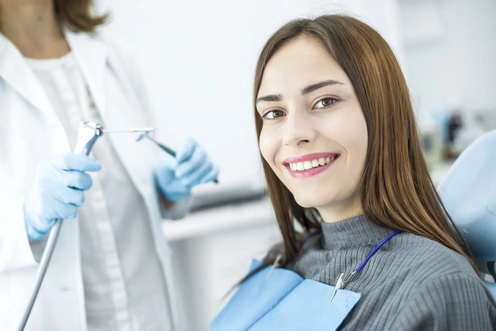 Smiling woman in dental chair with bib, dentist holding dental tools with gloves in background.