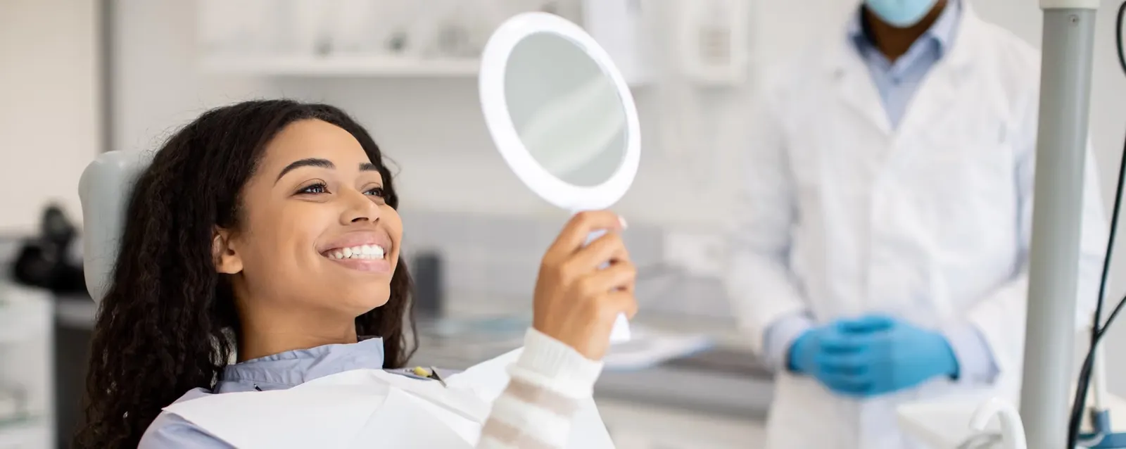 Smiling woman in dental chair holding mirror, dentist in white coat and gloves in background.
