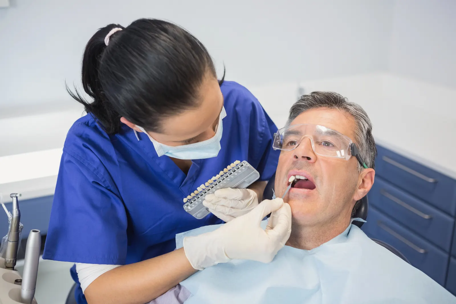 Dentist in blue scrubs matching dental veneers to a male patient's teeth in a dental clinic.