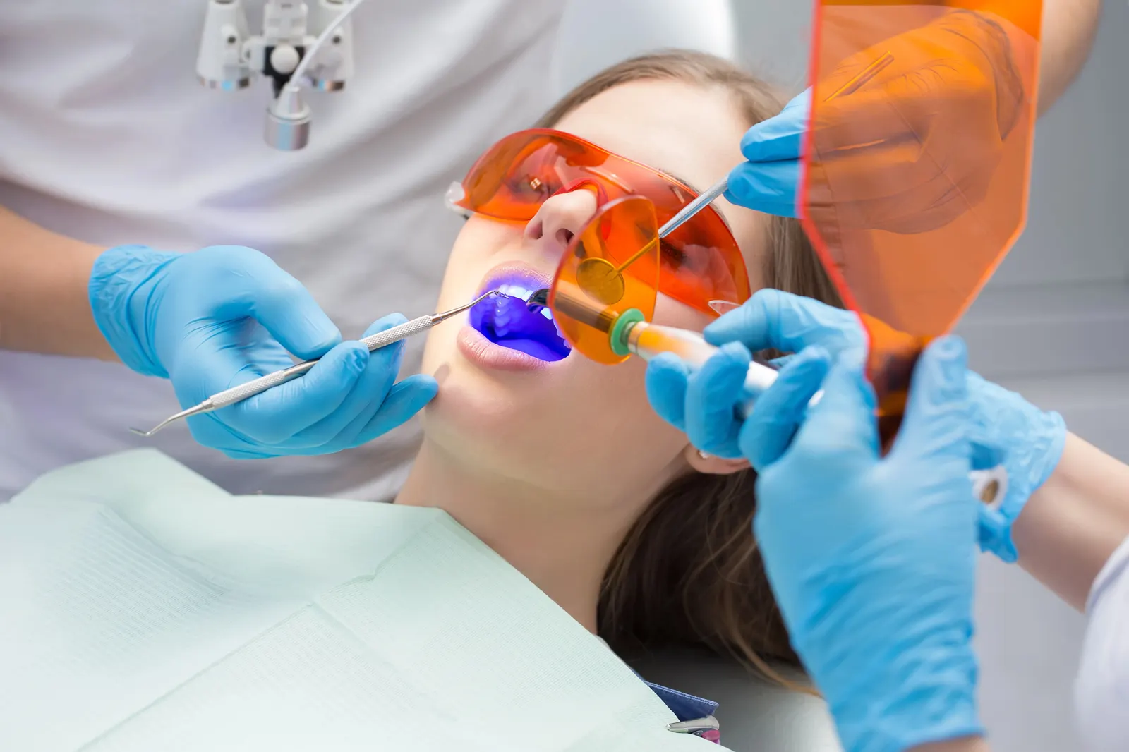 Dentist using tools and UV light to cure dental filling for patient wearing orange protective glasses.
