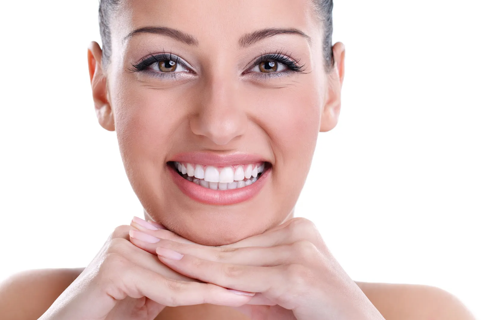 Close-up of a woman smiling with hands under chin showing white teeth and brown eyes on white background.