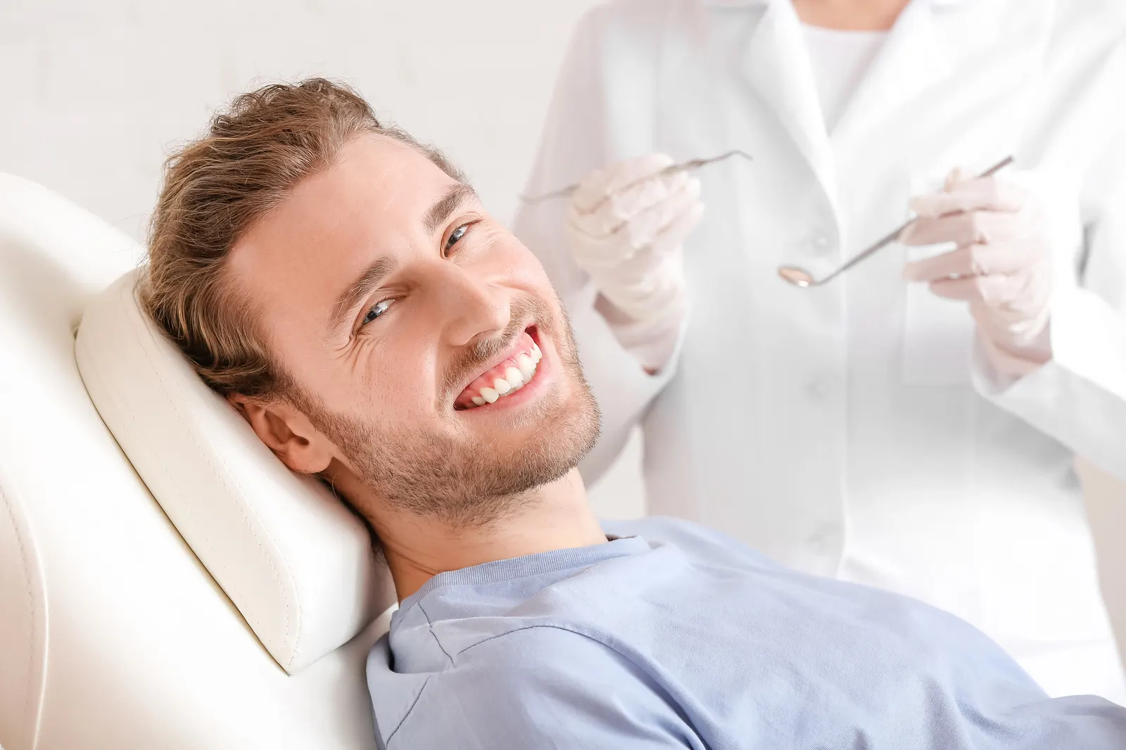 Smiling man reclining in a dental chair with a dentist holding dental tools in the background.