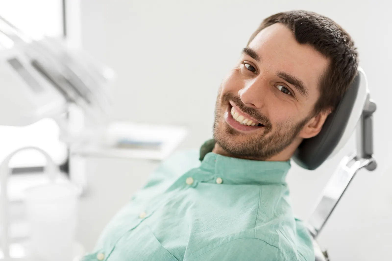 Smiling man in a teal shirt reclining in a dental chair at a dental office.