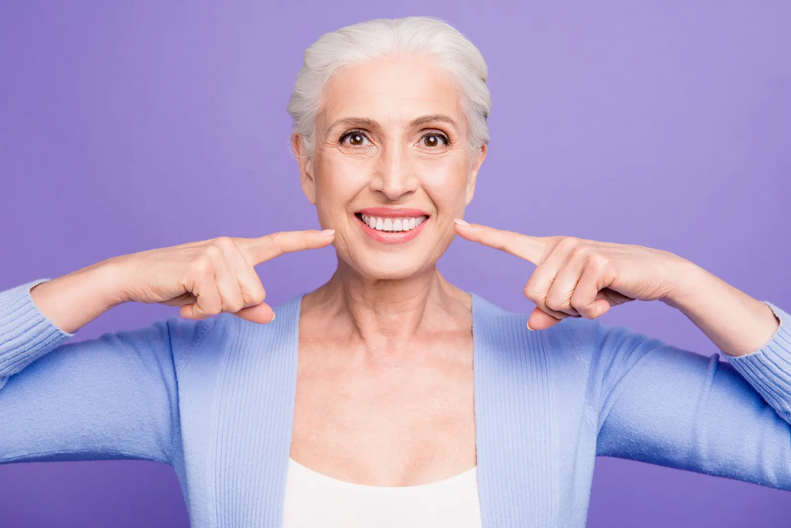 Older woman with white hair smiling and pointing to her teeth wearing a blue cardigan on purple background.