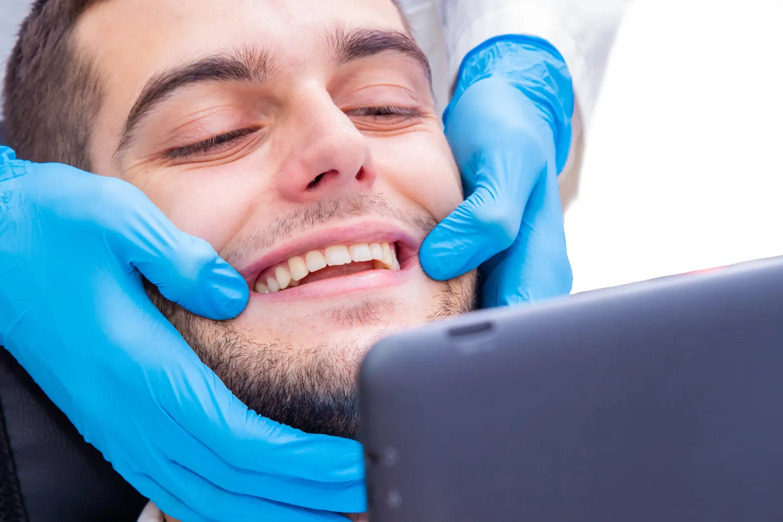 Close-up of a man smiling as gloved hands examine his teeth, with a tablet visible in the foreground.