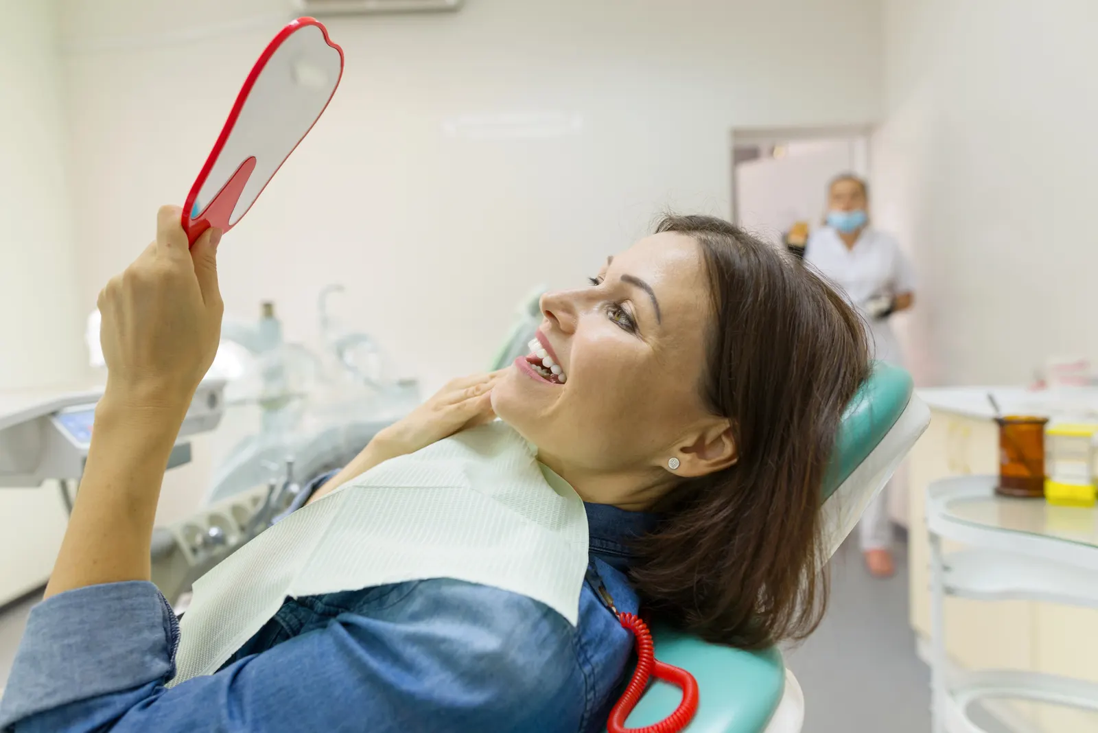 Woman in dental chair holding a hand mirror, smiling and inspecting her teeth after treatment.