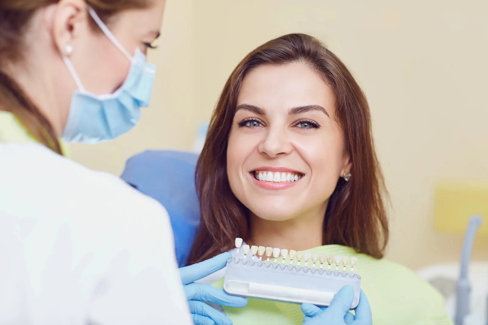 Dentist in gloves showing woman teeth shade guide as she smiles in dental chair.