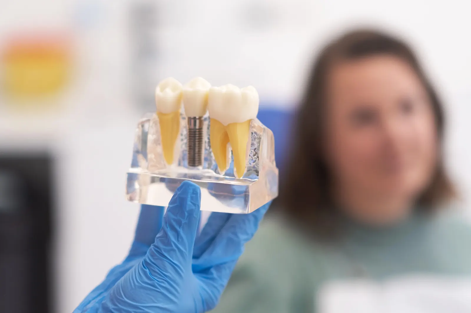 Gloved hand holding a dental model showing a natural tooth, implant, and crowned tooth.