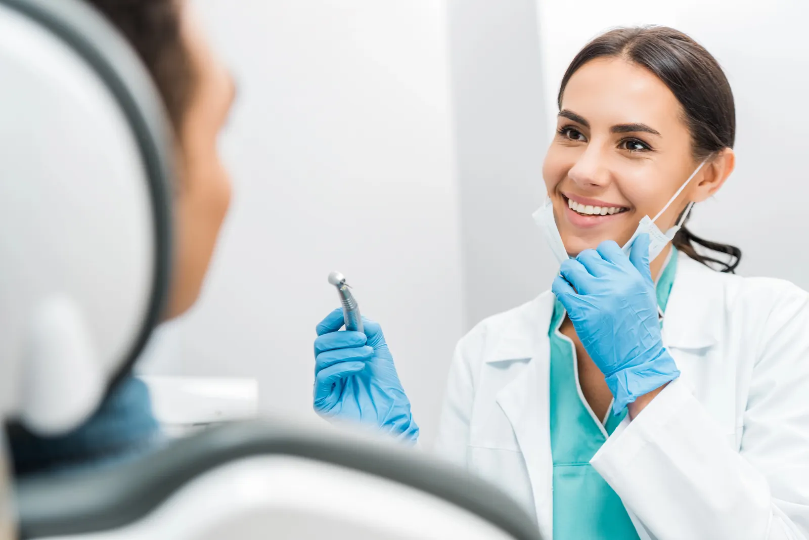 Smiling female dentist holding dental drill, adjusting her mask while talking with patient in clinic.