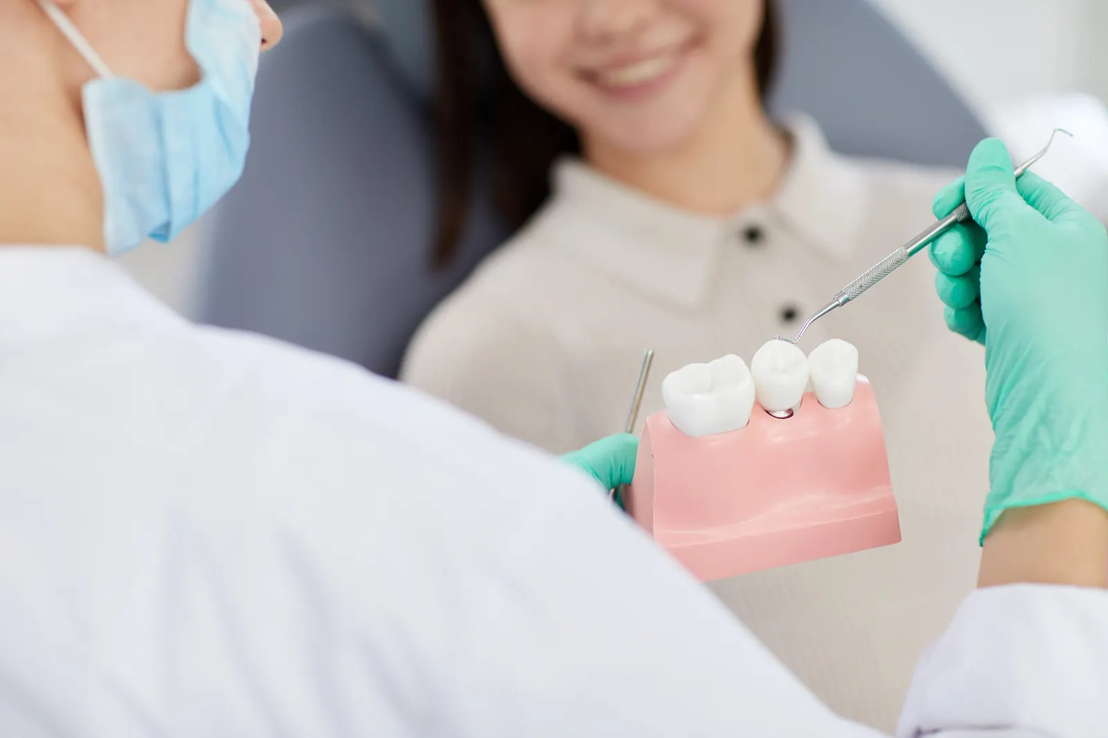Dentist wearing gloves and mask holds dental model explaining teeth to smiling patient.
