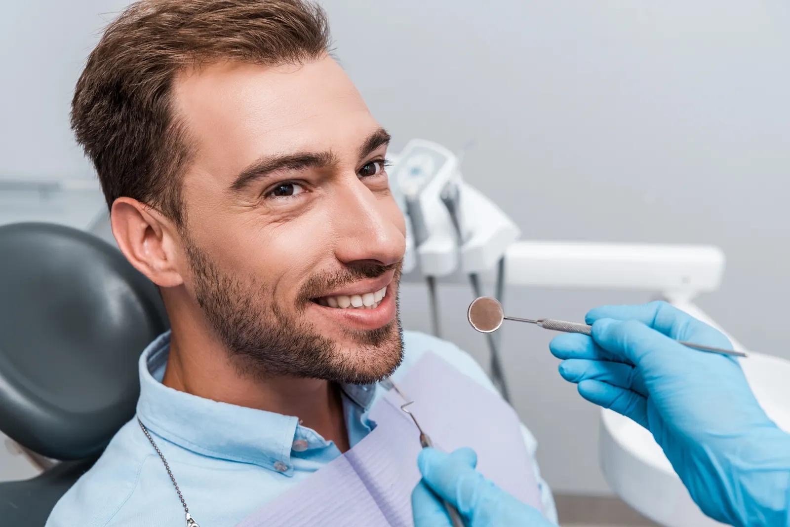 Man smiling in dental chair while a dentist wearing blue gloves holds dental tools near his mouth.