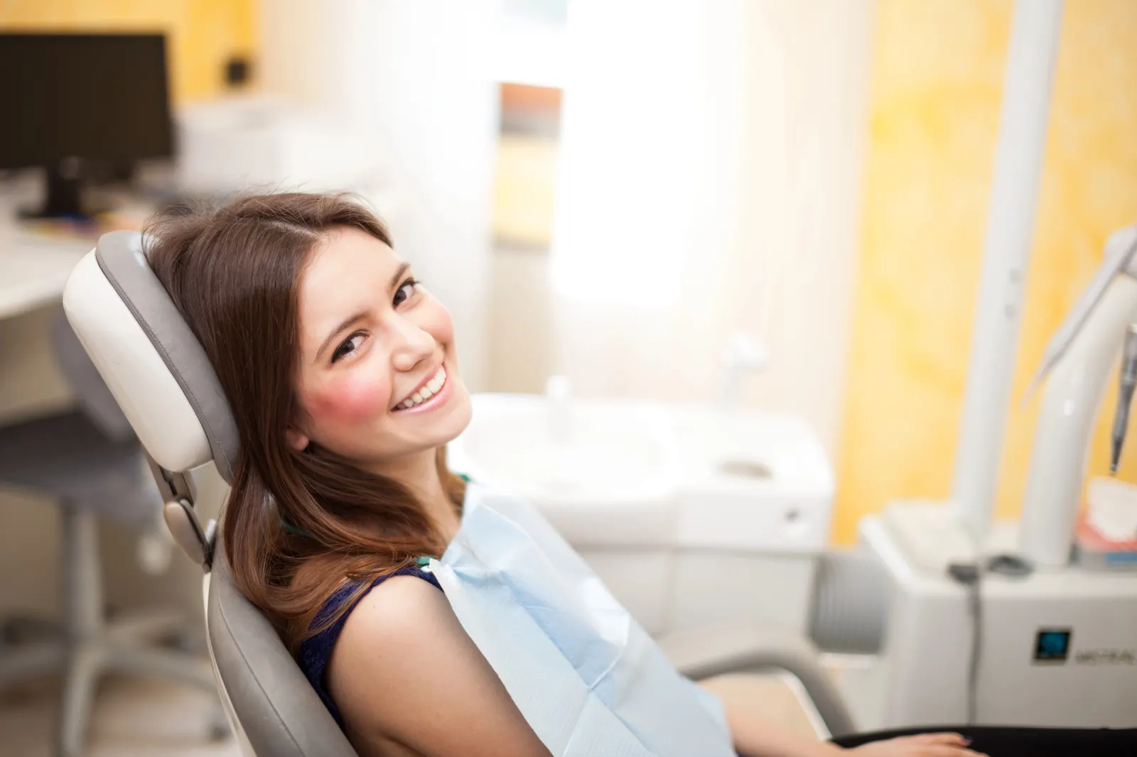 Smiling woman with long brown hair sitting in a dental chair wearing a dental bib.