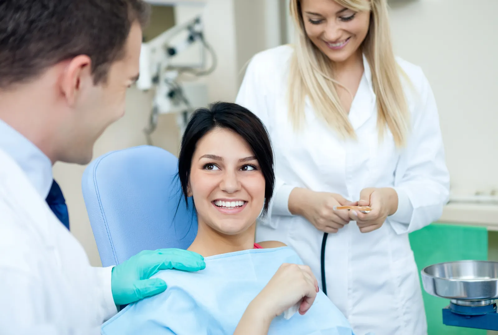 Smiling woman in dental chair talking to male dentist with female assistant nearby.