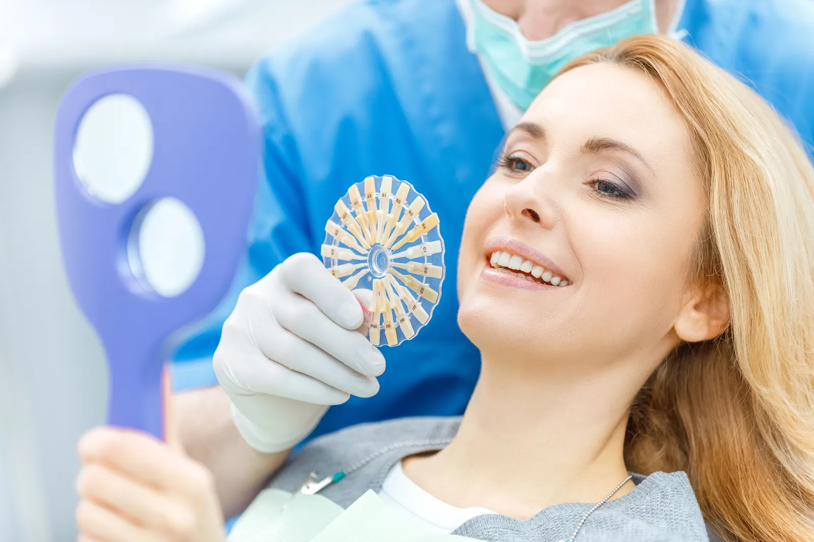 Dentist showing a woman shades of dental veneers while she smiles holding a mirror.